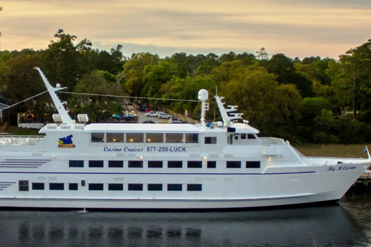 A casino cruise ship is pictured docked on calm waters, surrounded by lush green trees. The image conveys a sense of excitement and leisure, hinting at onboard dining, entertainment, and the unique opportunity to enjoy a sunset cruise along the coast.