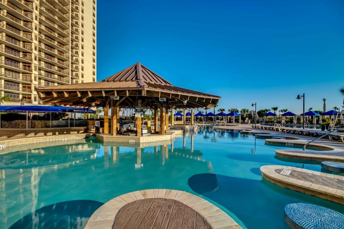An inviting pool area featuring a thatched-roof gazebo, loungers, palm trees, and blue umbrellas. Guests are swimming, sunbathing, socializing, and enjoying drinks poolside, creating a leisurely and vacation-like mood.