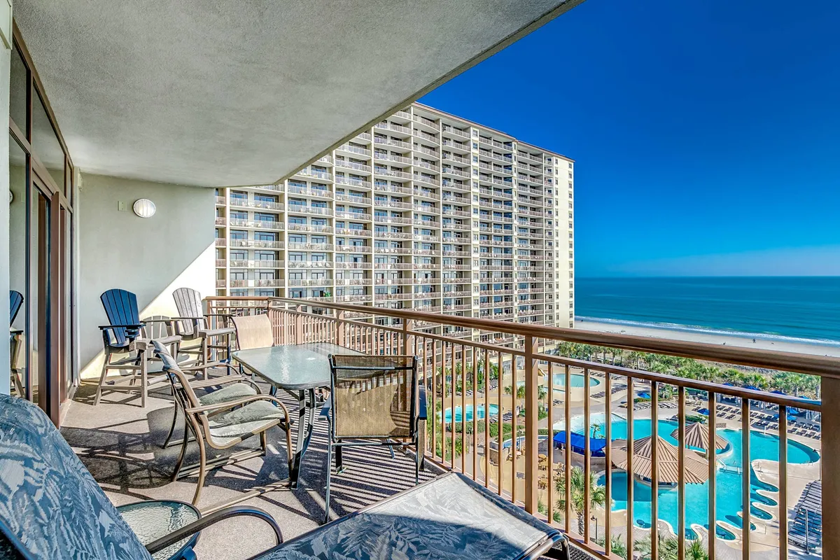 A serene exterior perspective from a private balcony, showing comfortable balcony furniture, a view over the swimming pool area, and the sparkling ocean beyond. The mood is peaceful and inviting, highlighting moments like enjoying morning coffee, reading a book, or watching the sunset.