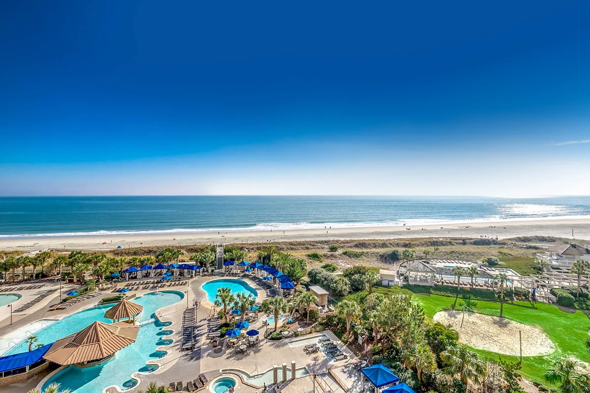 A vibrant exterior view of the North Beach Towers' expansive pool complex framed by lush palm trees, the sandy beach, and the ocean beyond. Families can be seen swimming, sunbathing, playing beach volleyball, and strolling along the shoreline, capturing the lively and relaxed atmosphere of the resort's outdoor amenities.