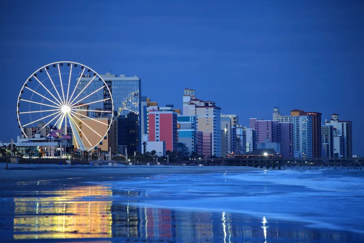 The image shows a lively nighttime scene featuring the iconic Myrtle Beach ferris wheel illuminated against the sky, with colorful buildings lining the oceanfront and the reflections shimmering on the water. The festive energy is palpable, evoking a sense of adventure and fun along the bustling boardwalk.
