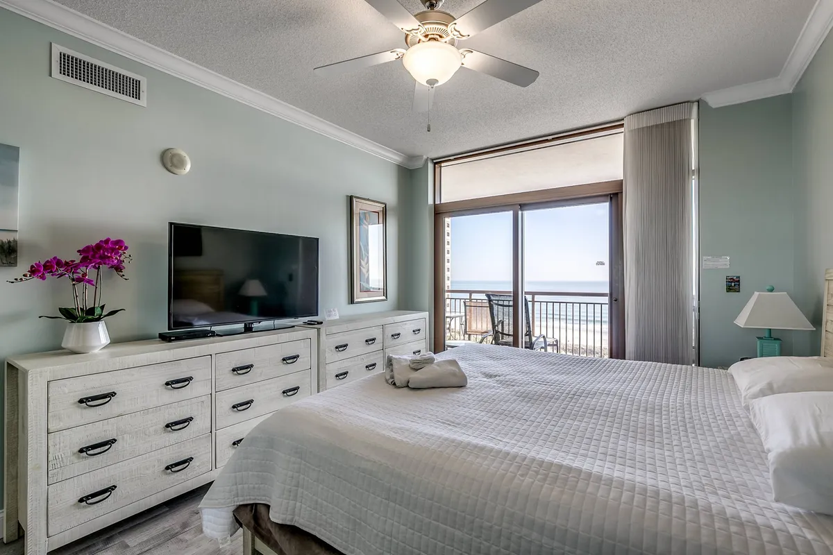 A spacious bedroom featuring a large bed with crisp white bedding, a dresser and television, and a window offering sweeping ocean views. The room is bathed in natural light, and the ceiling fan adds comfort. The ambiance is peaceful and serene, ideal for relaxation.