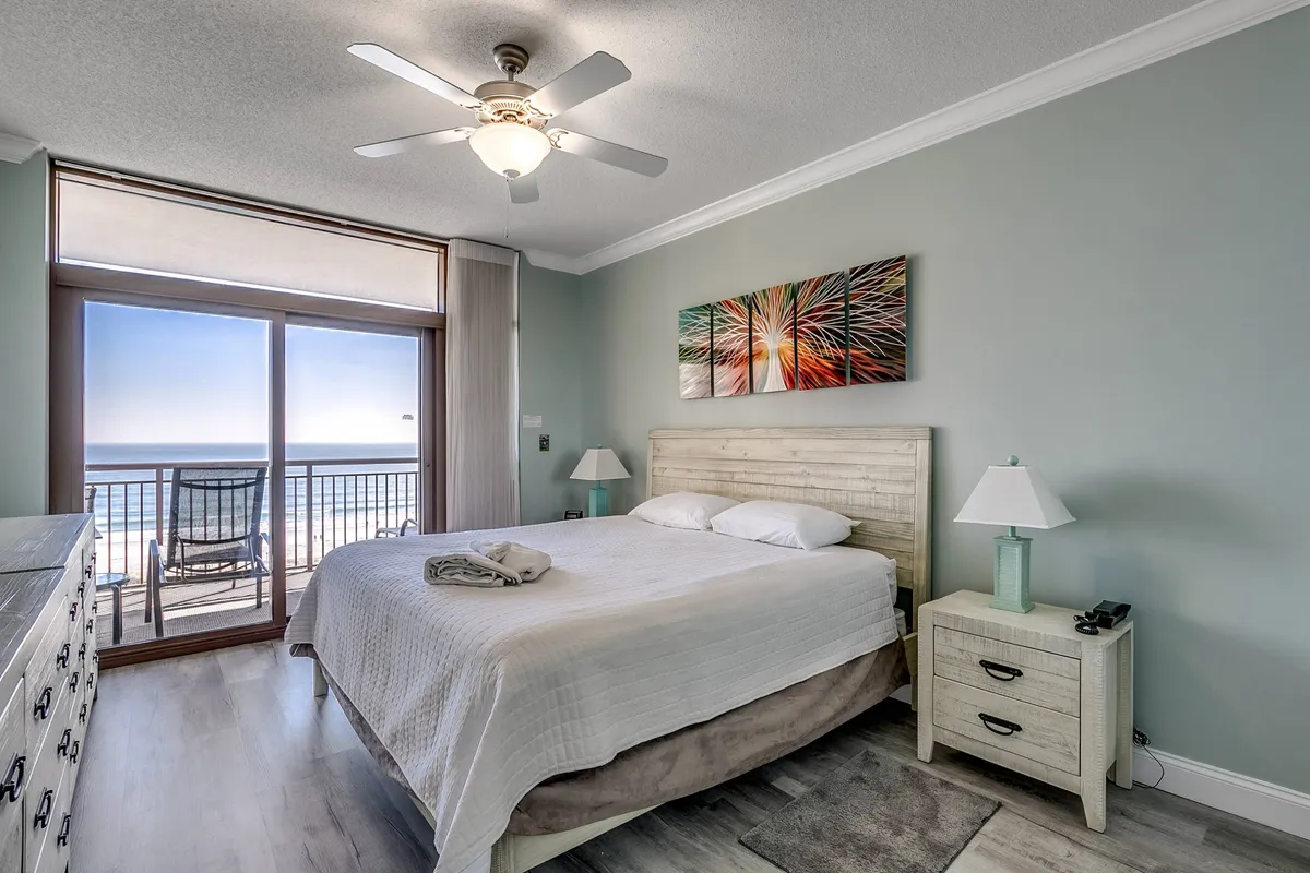 A spacious bedroom with a comfortable bed faces a large window, offering sweeping views of the ocean. Soft morning light pours in, highlighting cozy linens and tasteful artwork, creating a perfect space to unwind and take in the seaside scenery.