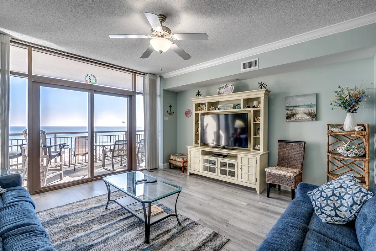 A bright, open living area featuring a comfortable sofa seating arrangement facing a large entertainment center with a flat-screen TV. Expansive windows offer sweeping views of the Atlantic, flooding the space with natural light and providing a direct connection to the oceanfront balcony. The overall atmosphere is casual and relaxed, ideal for family gatherings or laid-back evenings.