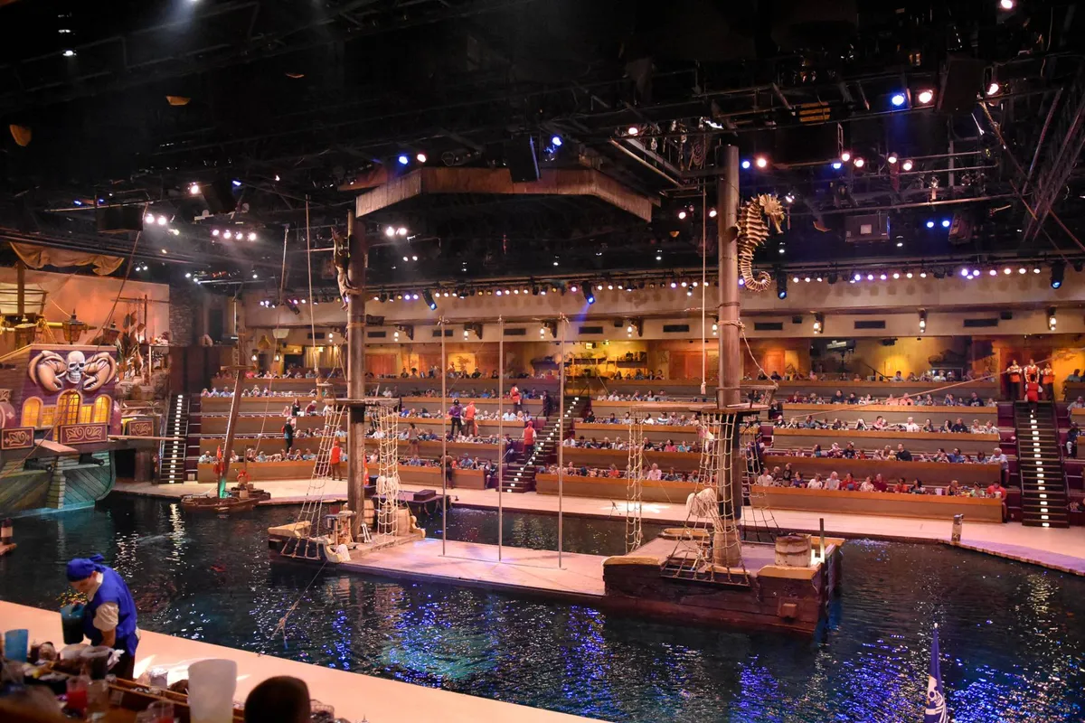 A lively interior shot of the Pirate’s Voyage Dinner & Show venue, featuring the dramatic stage set with a pirate ship and water feature, and an audience seated in anticipation of the interactive performance and themed dining experience.