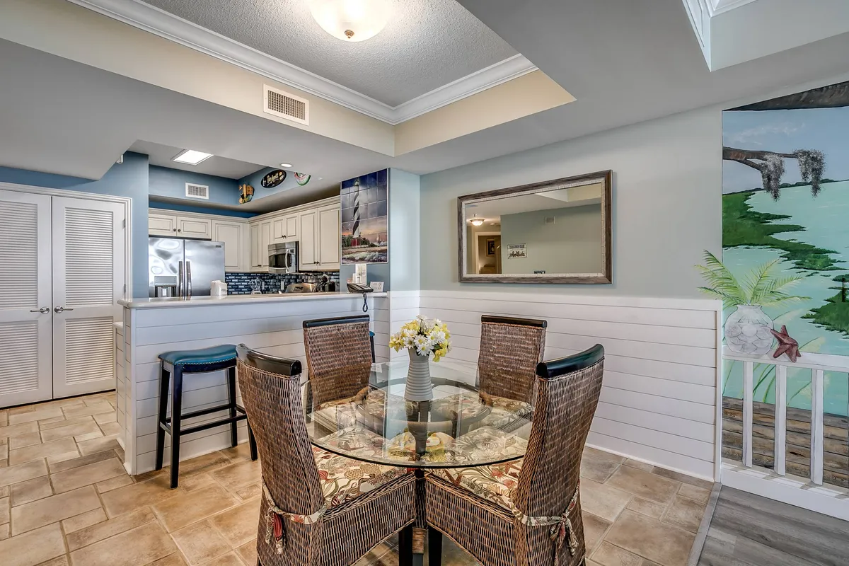 A warm, welcoming dining area with a modern glass dining table surrounded by woven chairs, set against a backdrop of tasteful wall art. The scene extends into a sleek kitchen, hinting at easy transitions between family meals, entertaining, and casual dining. Natural light and a cozy vibe encourage gathering and connection.