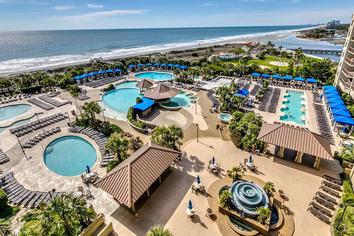 A vibrant exterior shot capturing multiple swimming pools bordered by palm trees, lounge chairs, and umbrellas, with the beach and ocean visible just beyond. Guests are swimming, sunbathing, relaxing by the pool, and walking along the beach, while the general mood is vacation-like and carefree.