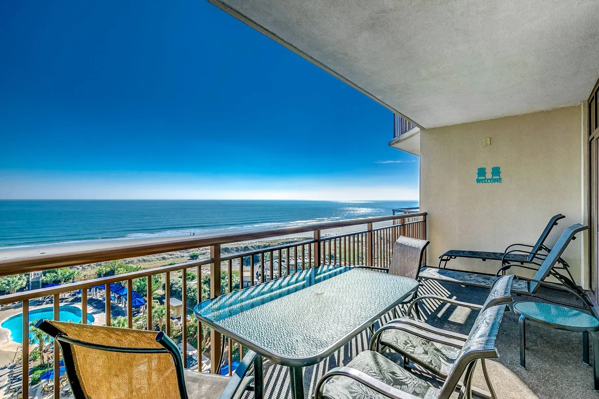 A tranquil covered balcony furnished for relaxation, with an uninterrupted view of the ocean and pool area below. This inviting space is ideal for unwinding after a sun-soaked day or gathering with loved ones to watch the sunset, embodying the seamless indoor-outdoor connection.