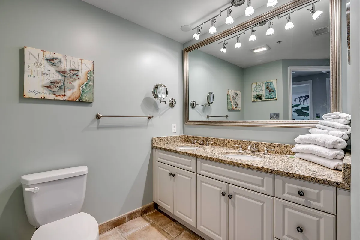 A bright and inviting bathroom scene with a granite countertop, large mirror, elegant towel display, and framed artwork. The setting feels luxurious and spa-like, offering a sense of rejuvenation and comfort.