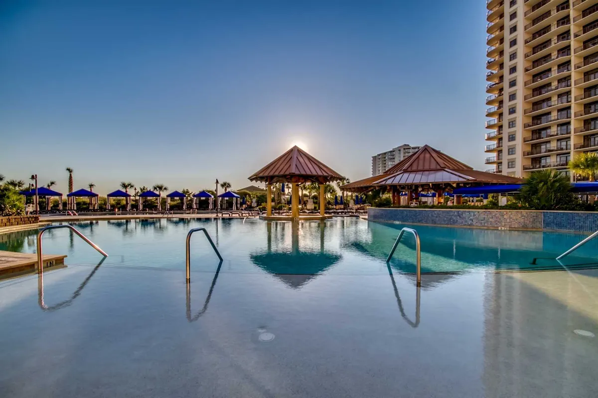 A sun-dappled resort pool bordered by thatched-roof cabanas, with the sun setting gracefully in the background. Guests lounge and socialize poolside, evoking post-golf relaxation and enjoyment just steps from the course.