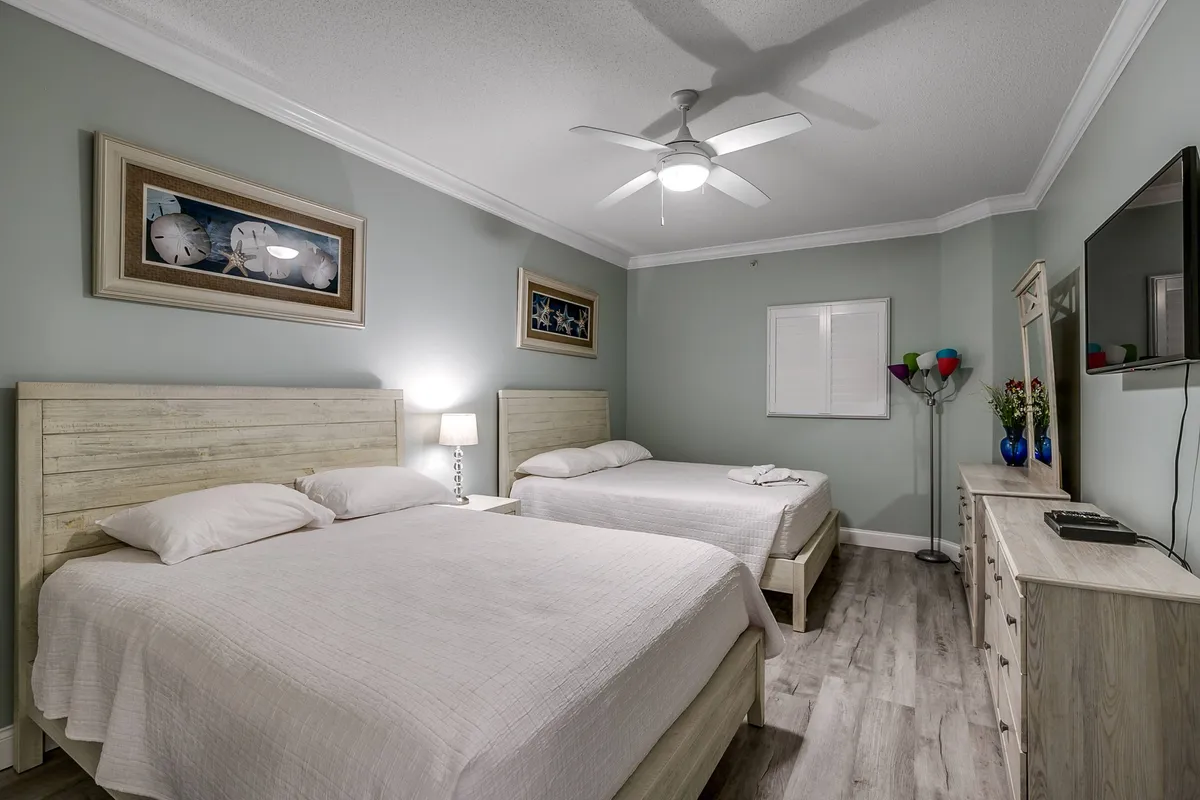 A comfortable bedroom with two queen beds, a central nightstand and lamp, a TV atop a dresser, and decorative artwork on the walls. The overall mood is calm and restful, inviting peaceful nights after an active day.