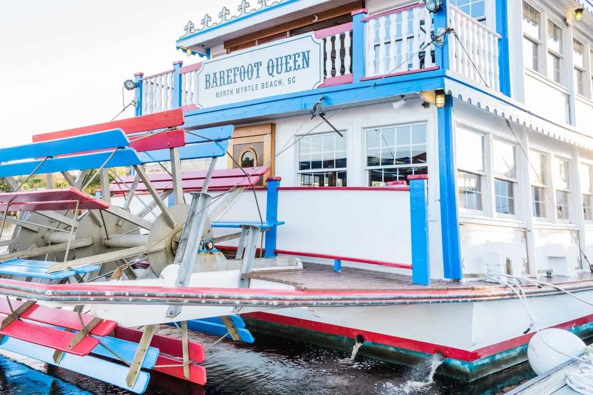 A vibrant paddle wheel boat with a colorful facade, lively signage, and guests mingling on the decks. The scene hints at festive dining and entertainment options nearby, tying into the narrative of exploring local hotspots after a day of golf.
