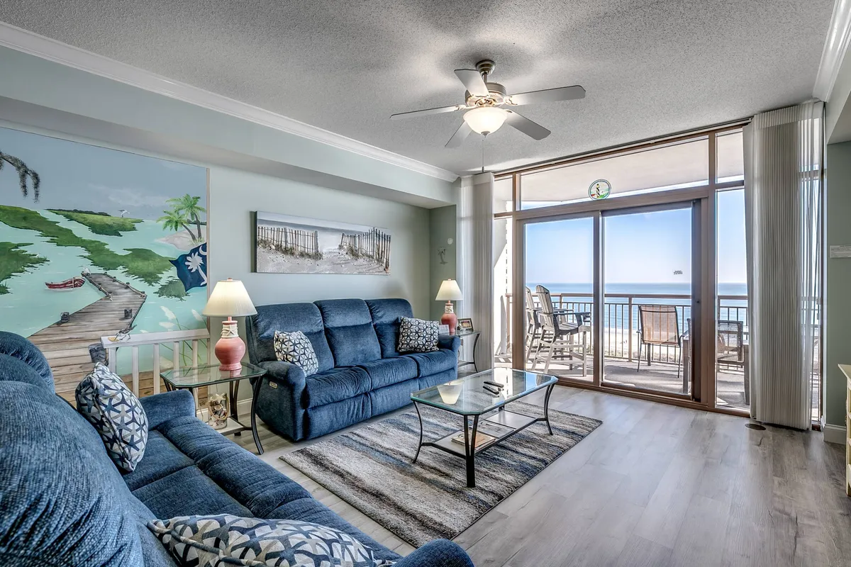 The image showcases a bright, spacious living area in a North Myrtle Beach condo, featuring plush blue couches arranged for conversation, striking coastal artwork on the walls, and expansive windows that frame breathtaking ocean views. Sunlight floods the room, and a glimpse of the balcony invites guests to step outside and bask in the sea breeze or watch the sunrise.