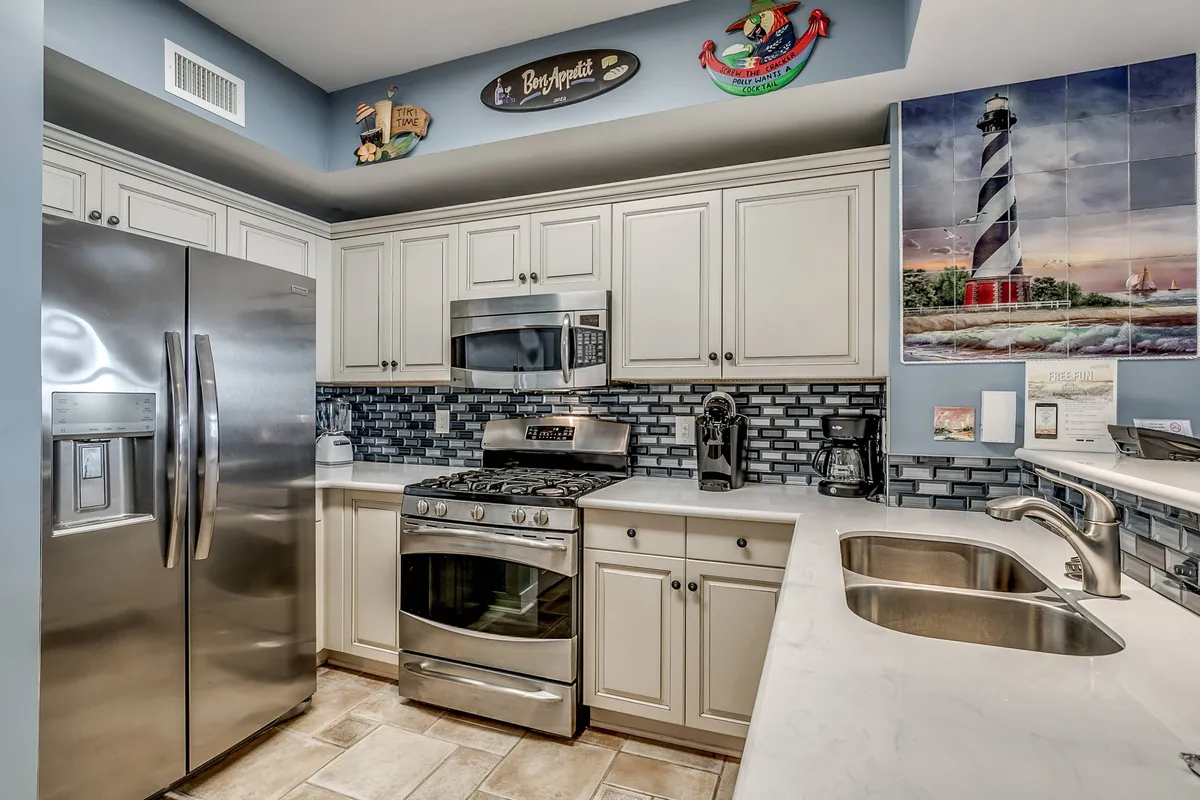 The image shows a welcoming kitchen featuring a stainless steel refrigerator, gas stove, a decorative lighthouse mural, and a blue backsplash. The space is laid out for both cooking and gathering, with ample counter space that makes it ideal for preparing meals and socializing.